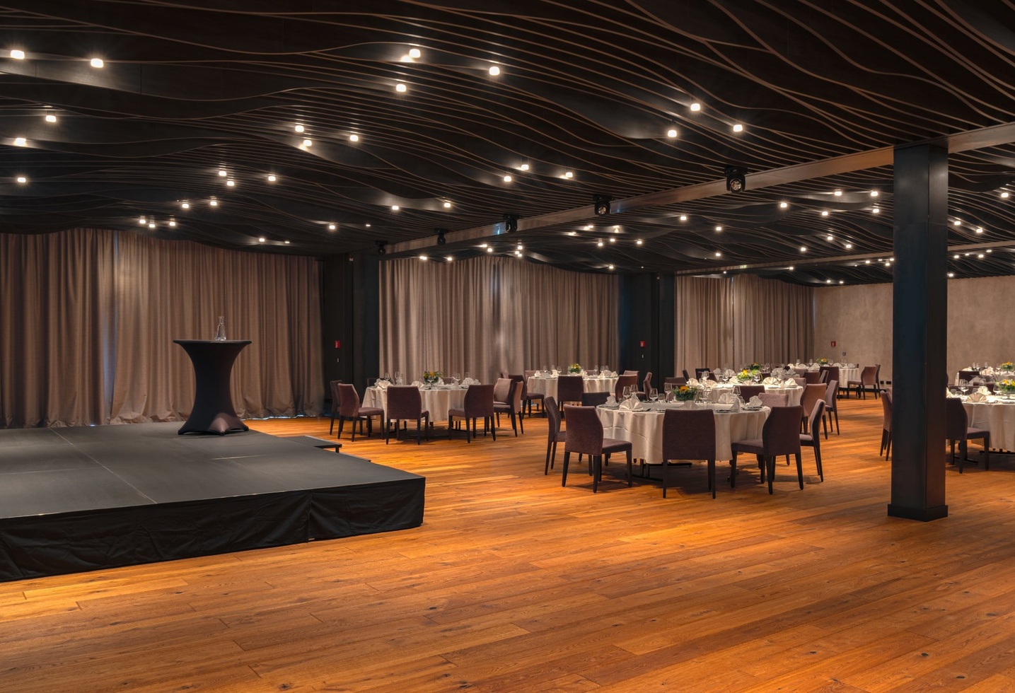 Eleganter Bankettsaal mit runden Tischen, Blumenschmuck, Holzboden und einer kleinen Bühne.