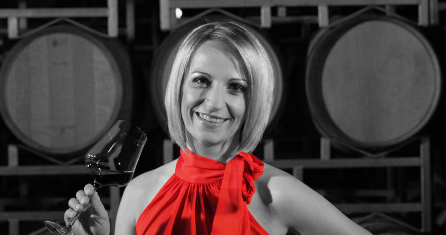 Woman in a red dress, smiling and holding a wine glass, standing in front of wine barrels.