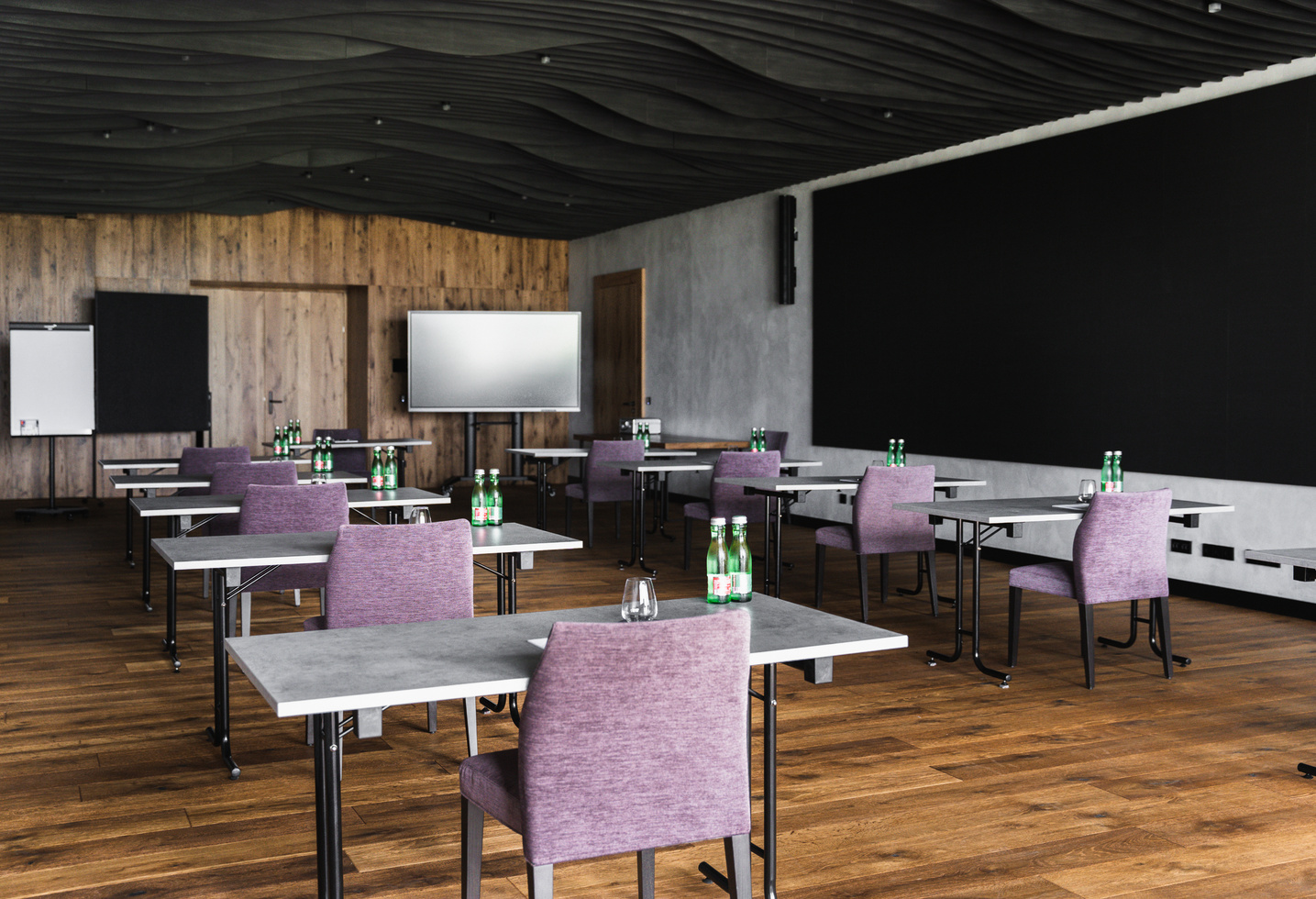Moderner Konferenzraum mit verteilten Tischen, violetten Stühlen, Wasserflaschen und Bildschirmen an der Stirnseite.
