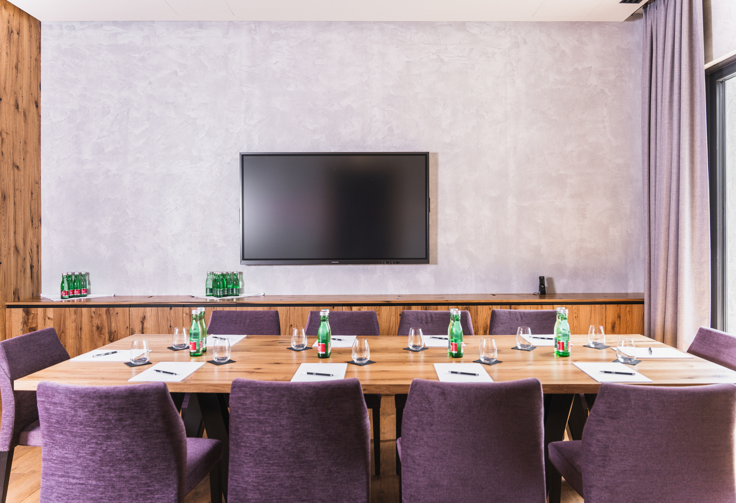 Konferenzraum mit großer Leinwand, Holztisch, lila Stühlen, Wasserflaschen und Notizblöcken.