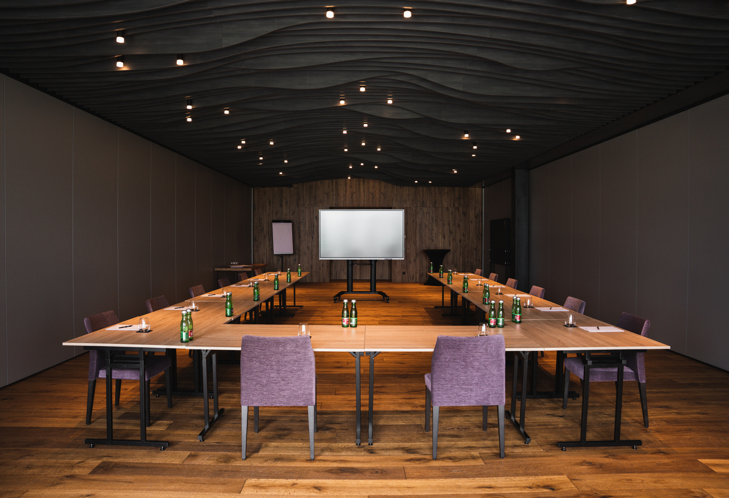 Moderner Konferenzraum mit U-förmigen Tischen, Stühlen, Wasserflaschen und einem leeren Bildschirm an der Stirnseite.