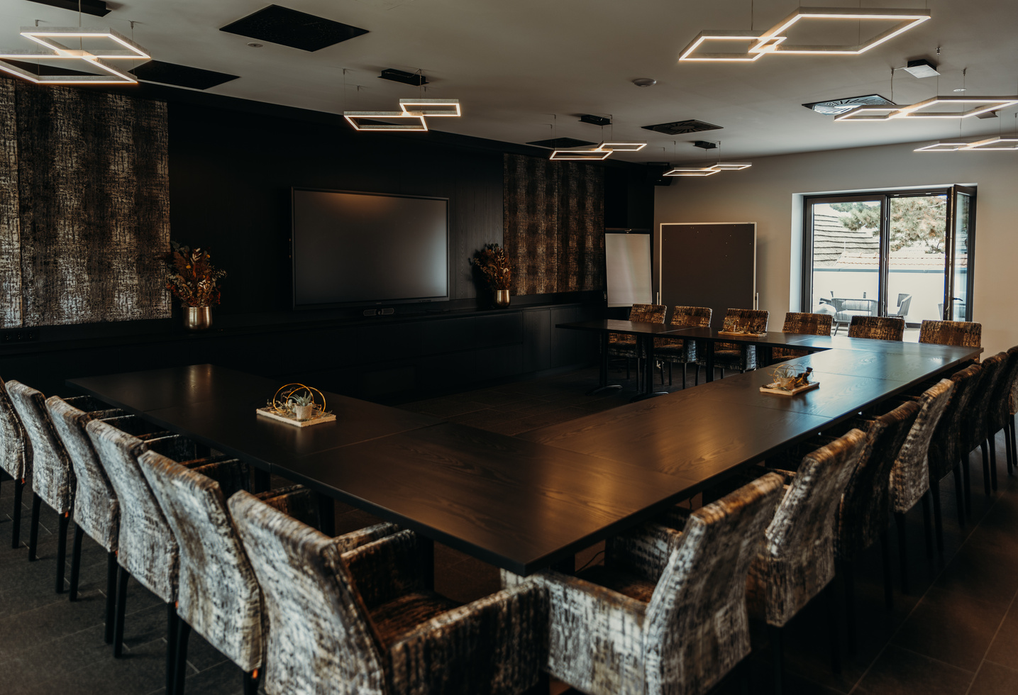 Moderner Konferenzraum mit U-förmigen schwarzen Tischen, gemusterten Stühlen und geometrischen Deckenleuchten.