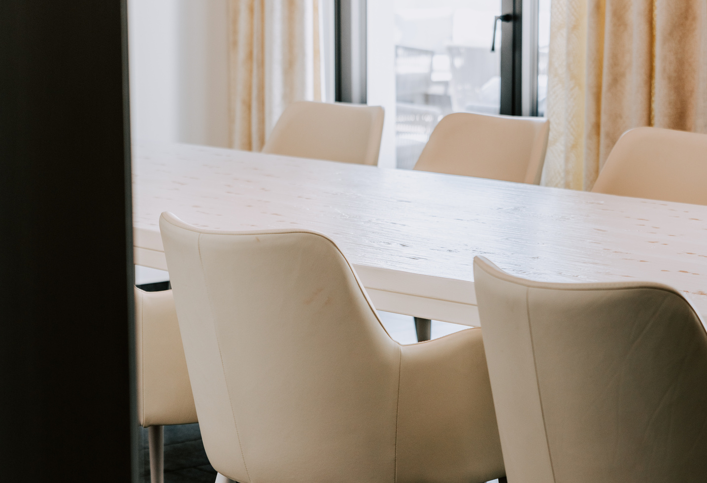 Weiße Stühle umgeben einen hellen Tisch in einem hellen Raum mit großen Fenstern und cremefarbenen Vorhängen.