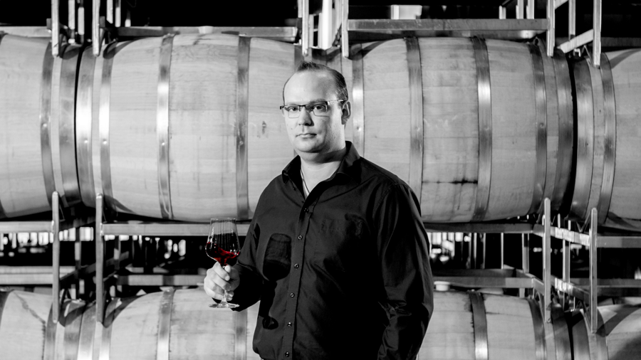 Ein Mann mit Brille hält ein Glas Rotwein in der Hand und steht vor gestapelten Weinfässern aus Holz.