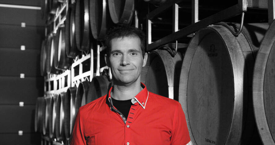 A man in a red shirt stands smiling in front of stacked wine barrels in a cellar.