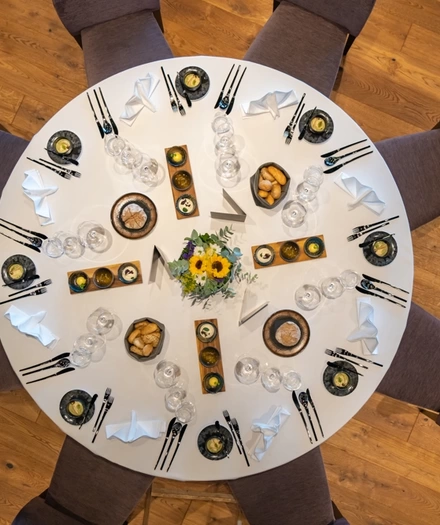 Ein runder, für acht Personen gedeckter Tisch mit Tellern, Gläsern, Besteck, Servietten und einem floralen Tafelaufsatz, von oben gesehen.