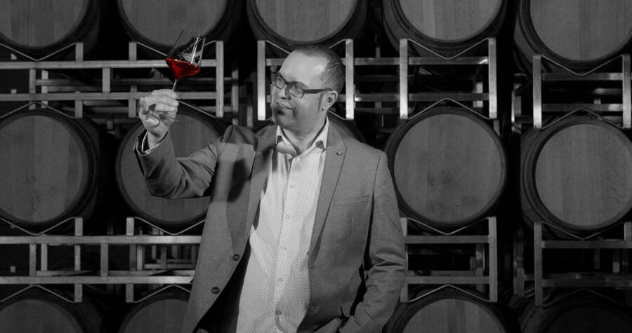 Ein Mann im Anzug prüft ein Glas Rotwein vor gestapelten Weinfässern in einer Weinkellerei.