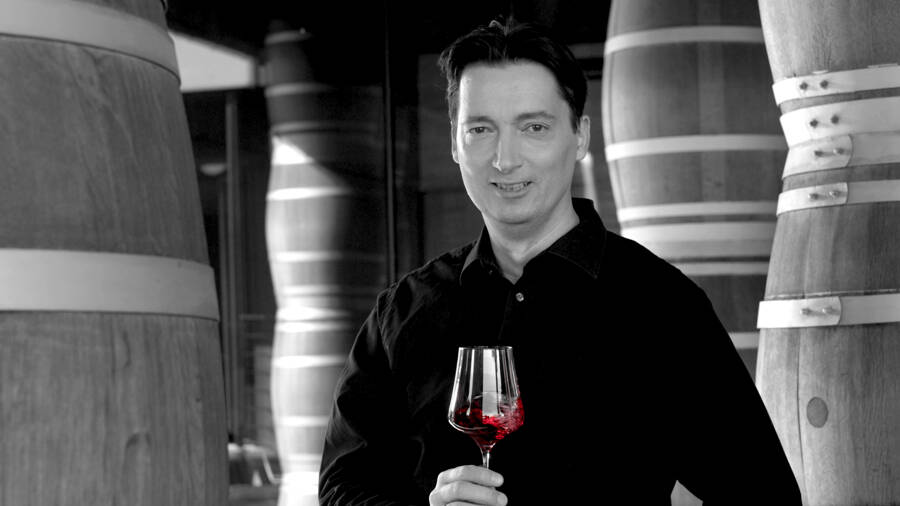 A man with a glass of red wine in his hand stands between large wooden barrels in a wine cellar.