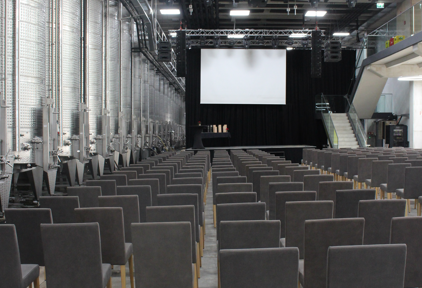 In einem modernen industriellen Veranstaltungsraum stehen leere Stuhlreihen vor einer Bühne mit einer leeren Leinwand.