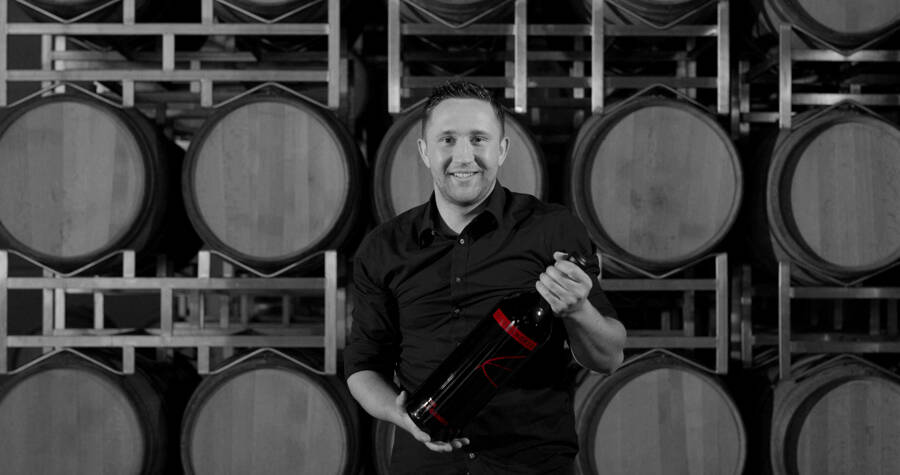 Ein lächelnder Mann mit einer großen Weinflasche in der Hand steht vor gestapelten Weinfässern in einer Weinkellerei.
