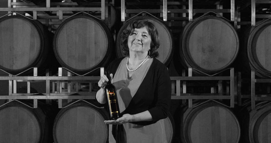 Frau mit einer Weinflasche in der Hand vor gestapelten Holzfässern in einer Weinkellerei.