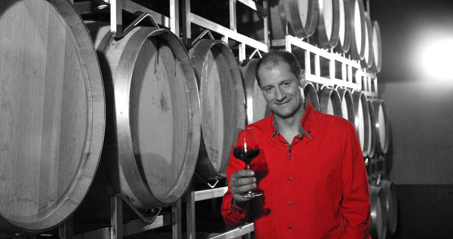 Ein Mann in einem roten Hemd hält ein Glas Rotwein in der Hand und steht neben gestapelten Weinfässern in einem Keller.