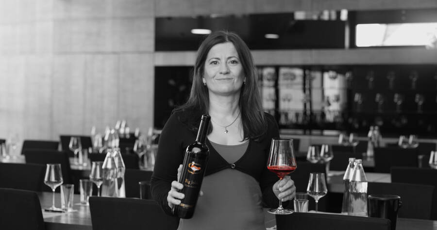 Lächelnde Frau in einem Restaurant, die eine Weinflasche und ein Glas Rotwein in der Hand hält; gedeckte Tische im Hintergrund.