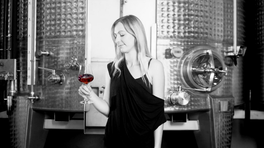 Eine Frau in einem schwarzen Kleid hält ein Glas Rotwein vor großen metallenen Weintanks.