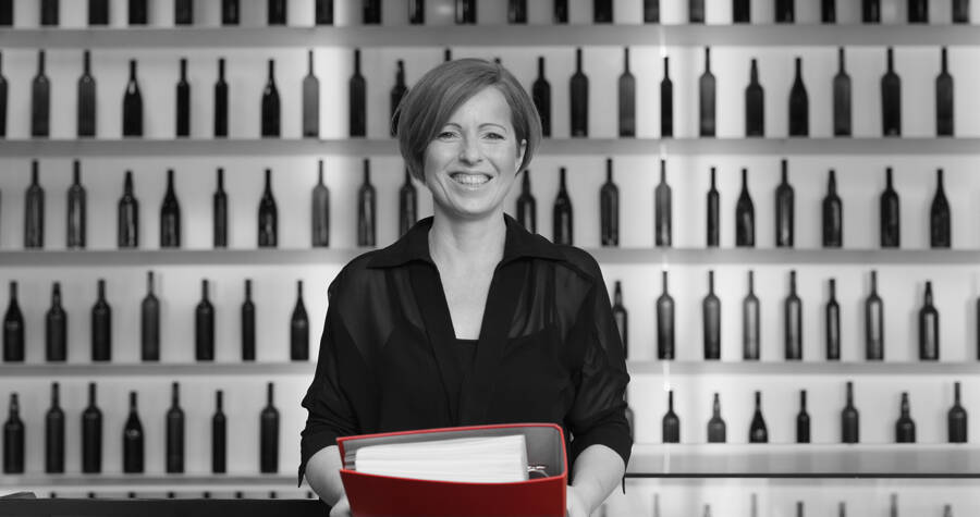 Lächelnde Person, die einen roten Ordner in der Hand hält und vor einem Regal mit Weinflaschen steht.