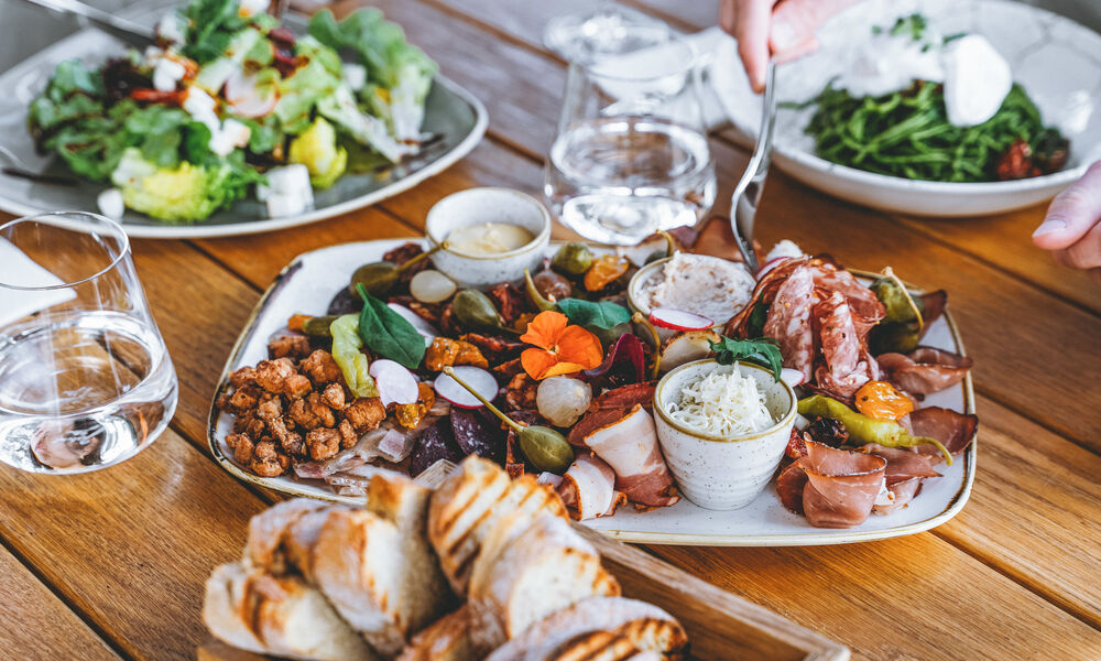Eine Wurstplatte mit Wurst, Käse, Oliven, Brot und Dips auf einem Holztisch, auf dem eine Hand das Essen serviert.