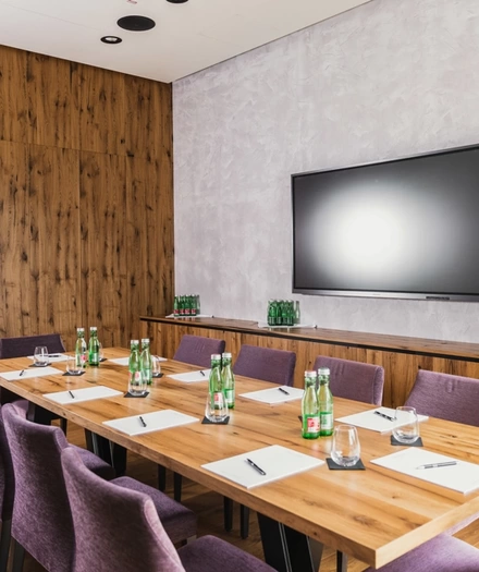 Modern conference room with wooden table, purple chairs, notepads, pens and bottles of water. Television on the wall.