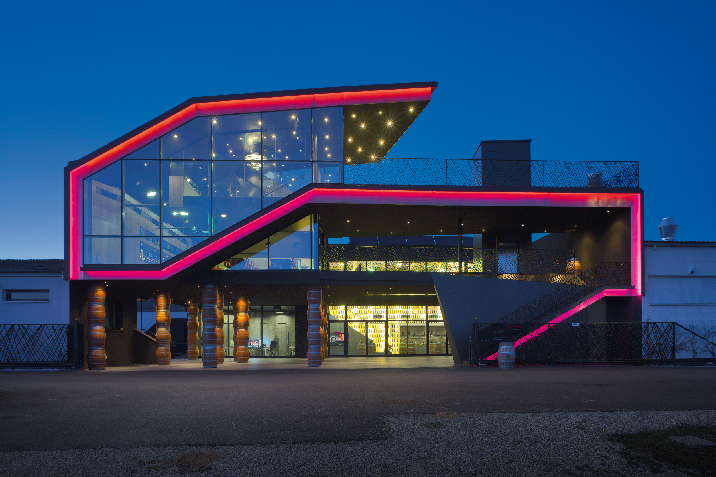 Modernes Gebäude mit großen Glasfenstern und rosafarbenen Neonlichtern, die in der Abenddämmerung das kantige Dach und die Treppe hervorheben.