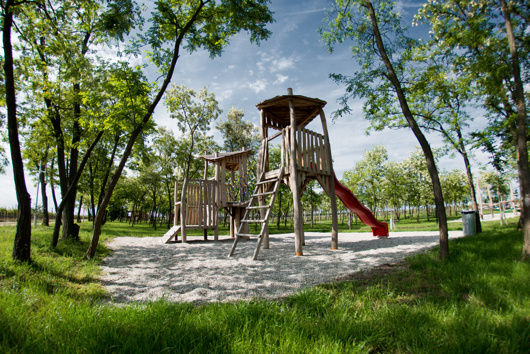 Spielplatz aus Holz mit Rutsche und Klettermöglichkeiten, umgeben von Bäumen und Gras an einem sonnigen Tag.