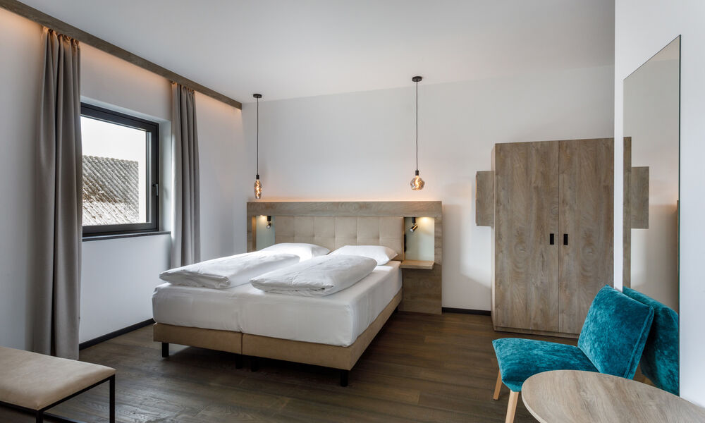 Modernes Hotelzimmer mit einem Doppelbett, Holzmöbeln, Hängelampen und einem blaugrünen Stuhl am Fenster.
