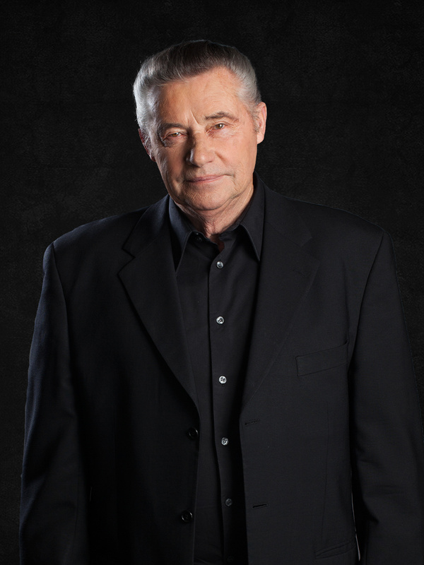 Ein älterer Mann mit grauem Haar in schwarzem Anzug und Hemd steht vor einem dunklen Hintergrund und schaut in die Kamera.